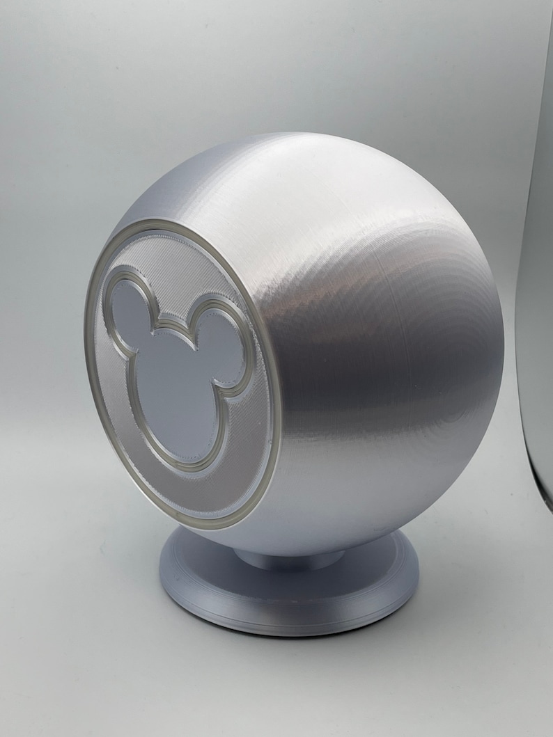 May include: A silver sphere-shaped decorative object with a Mickey Mouse silhouette on the front. The object is mounted on a circular base. The design features a textured surface and a smooth, metallic finish.