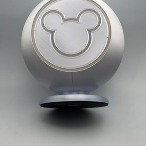 May include: A silver, spherical decorative object featuring a prominent Mickey Mouse silhouette. The object has a circular base and a smooth, metallic finish. The Mickey Mouse design is etched into the surface.