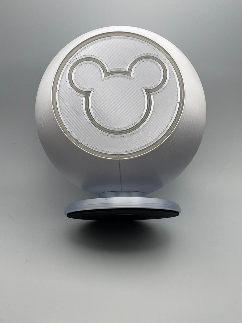 May include: A silver, spherical decorative object featuring a prominent Mickey Mouse silhouette. The object has a circular base and a smooth, metallic finish. The Mickey Mouse design is etched into the surface.