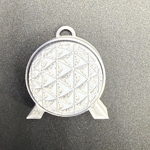 May include: A silver-colored pendant featuring a geometric sphere design with a loop at the top for hanging. The sphere has a textured surface with a pattern of interconnected triangles, and is supported by two small, angled feet.
