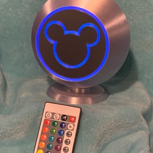 May include: A silver-colored, spherical lamp with a blue illuminated outline of a Mickey Mouse head. A remote control with color options is in front of the lamp. The lamp sits on a small, round base.