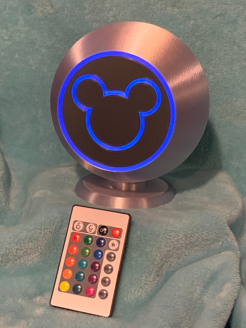 May include: A silver-colored, spherical lamp with a blue illuminated outline of a Mickey Mouse head. A remote control with color options is in front of the lamp. The lamp sits on a small, round base.