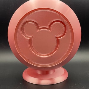 May include: A rose-colored, 3D-printed decorative stand featuring a circular design with a Mickey Mouse silhouette in the center. The stand has a round base and a textured surface, suitable for display.