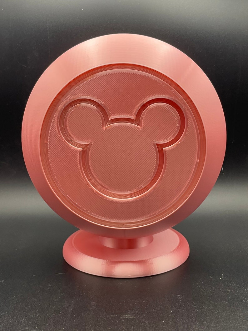 May include: A rose-colored, 3D-printed decorative stand featuring a circular design with a Mickey Mouse silhouette in the center. The stand has a round base and a textured surface, suitable for display.