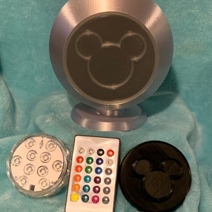 May include: A silver-colored, spherical lamp with a black Mickey Mouse silhouette on the front. Includes a remote control, a clear light base with multiple LEDs, and a black disc with a Mickey Mouse silhouette.