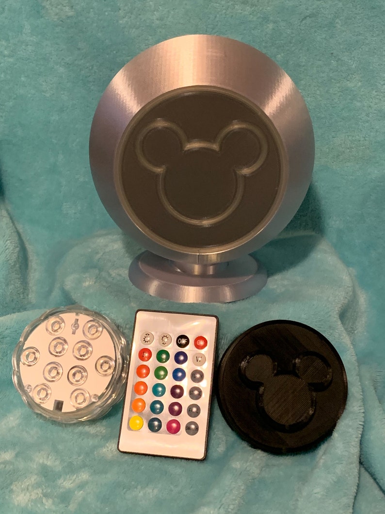 May include: A silver-colored, spherical lamp with a black Mickey Mouse silhouette on the front. Includes a remote control, a clear light base with multiple LEDs, and a black disc with a Mickey Mouse silhouette.