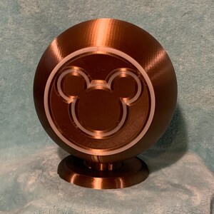 May include: A brown sphere-shaped decorative object with a Mickey Mouse silhouette design. The object is mounted on a small, circular base. The design is a light color, and the object has a textured surface.