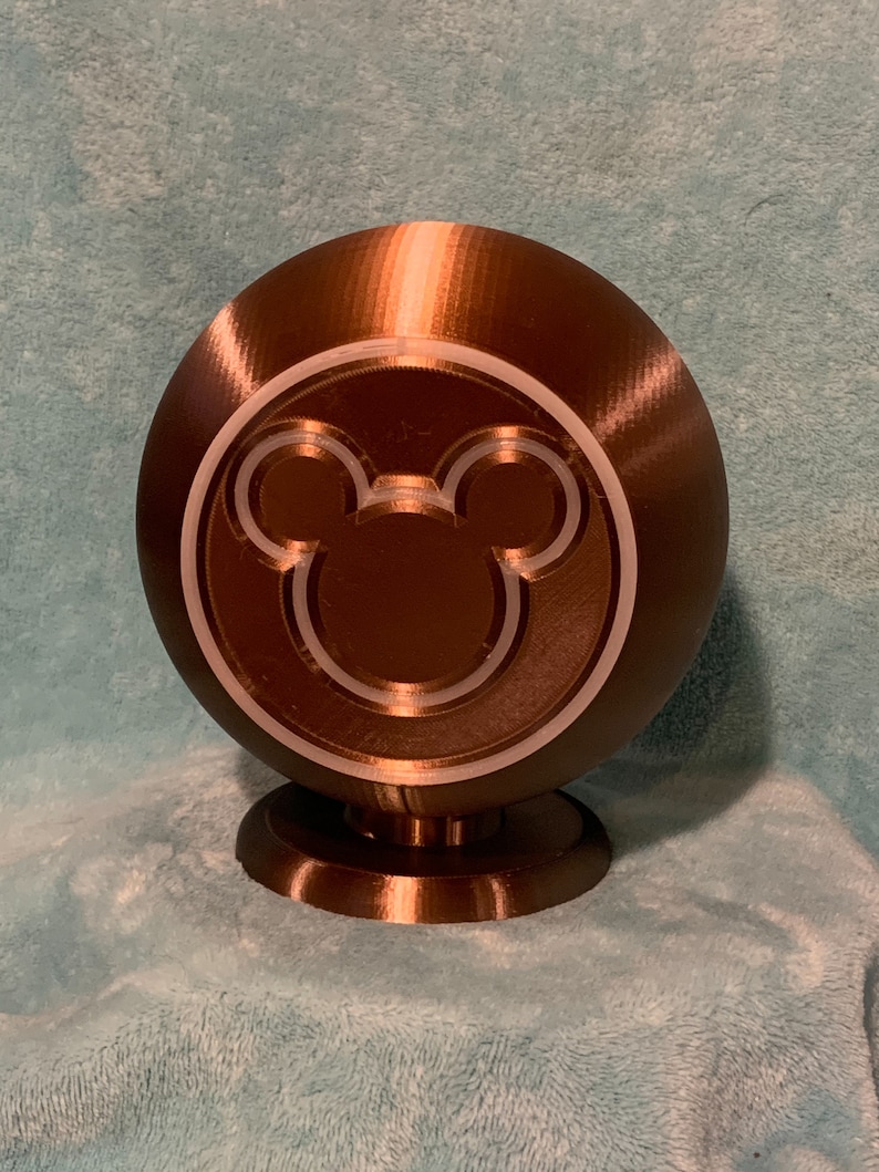 May include: A brown sphere-shaped decorative object with a Mickey Mouse silhouette design. The object is mounted on a small, circular base. The design is a light color, and the object has a textured surface.
