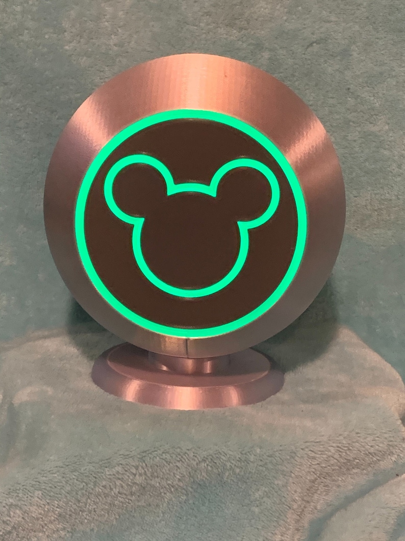 May include: A silver-colored, circular decorative object with a green glowing outline of a Mickey Mouse head. The object has a dark brown center and sits on a small, matching silver base. The design is simple and modern.