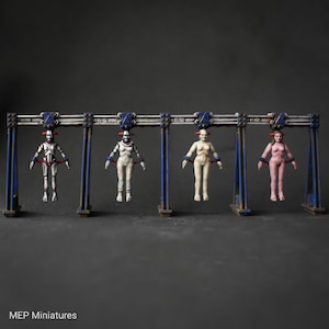 Könnte beinhalten: Vier Figuren hängen an einem Metallrahmen. Die Figuren sind in verschiedenen Farben bemalt, darunter Weiß, Pink und ein metallisches Silber. Die Figuren tragen Fesseln an Handgelenken und Knöcheln. Der Rahmen ist blau und weiß gestrichen mit den Buchstaben "EW" auf jeder Seite.