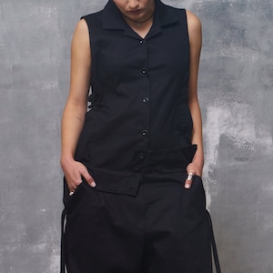 Könnte beinhalten: Ein schwarzes ärmelloses Hemd mit Knopfleiste und eine passende, weite Hose mit langen Bändern an den Seiten. Das Outfit wird von einer Person mit langen braunen Haaren getragen.
