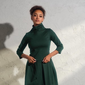 Peut inclure: Une femme portant un haut à col roulé vert foncé et une jupe vert foncé assortie. La jupe est plissée et présente un design asymétrique fluide. Elle porte des bottines noires.