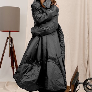 Può includere: Lungo cappotto trapuntato nero con ampio colletto. Il cappotto è lungo fino al pavimento e ha una silhouette voluminosa. La modella indossa stivali neri. Una lampada da terra a treppiede in legno è sullo sfondo.
