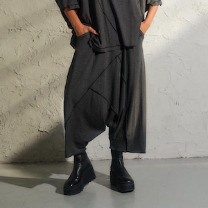 Puede incluir: Una mujer lleva una blusa y pantalones grises holgados y asimétricos. La blusa y los pantalones tienen un diseño geométrico con costuras negras. Lleva botines negros.