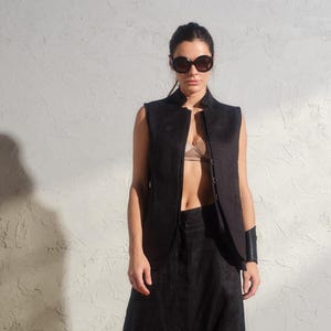 May include: A woman wearing a black sleeveless vest with a black bra underneath, black pants, and black heeled sandals. She is wearing sunglasses and a black bracelet on her left wrist.
