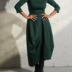 May include: A woman wearing a dark green turtleneck top and a long, flowing dark green skirt. She is standing in front of a white textured wall.