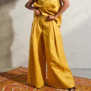 May include: A woman wearing a yellow linen sleeveless top and matching wide-leg pants. She is wearing round sunglasses and has curly hair.
