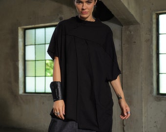 May include: A person wearing a black, loose-fitting shirt with a long, asymmetrical hemline and dark denim pants with a unique, gathered design. They are wearing gray sneakers.