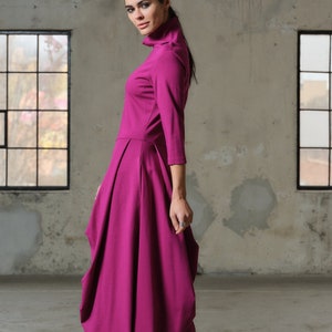 Peut inclure: Une femme portant une robe longue à manches longues magenta avec un col montant et une jupe plissée. Elle porte des bottes noires et se tient dans une pièce aux murs en béton et des fenêtres.