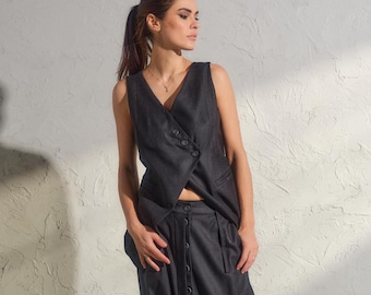 Op de afbeelding: Een donkergrijze mouwloze vest met een asymmetrische knoopsluiting, gecombineerd met een bijpassende broek met laag kruis. De broek heeft een knoopsluiting en zakken. De outfit wordt gecompleteerd met zwarte enkellaarsjes. De stof is getextureerd.