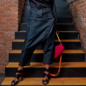 Op de afbeelding: Een persoon draagt een zwart overhemd met knopen en een donkerblauwe jeansbroek met wikkelstijl. De outfit is gecombineerd met zwarte sandalen, een roze handtas met een oranje band en een blauw getinte bril. De achtergrond is een bakstenen muur en een trap.