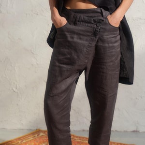 Pode incluir: Uma calça de linho cinza escuro com um design assimétrico único e fecho de botão. A calça é cropped e tem um caimento relaxado.