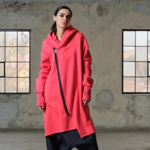 May include: A long, pink hooded sweatshirt with a diagonal zipper. The sweatshirt has a relaxed fit and a slightly asymmetrical hem. The model is wearing the sweatshirt with black pants and black platform boots.