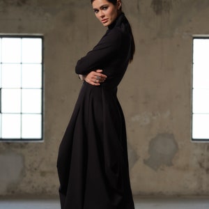 Peut inclure: Une femme portant une robe noire à manches longues avec une jupe plissée et des bottes à plateforme noires. Elle se tient dans une pièce aux murs en béton et aux fenêtres.