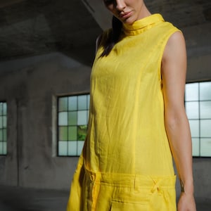 May include: A sleeveless, yellow jumpsuit with a high collar and a relaxed fit. The jumpsuit features a button closure, pockets, and decorative ties at the waist. The garment is made of a lightweight fabric.