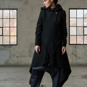 Puede incluir: Una chaqueta negra con capucha y un largo dobladillo asimétrico. La chaqueta tiene una cremallera y un cuello alto. La modelo lleva la chaqueta con botas de plataforma negras y pantalones azul oscuro.
