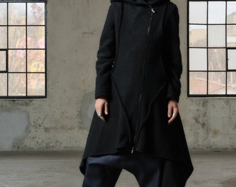May include: A black hooded coat with a long, asymmetrical hemline. The coat has a zipper closure and a high collar. The model is wearing the coat with black platform boots and dark blue pants.