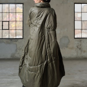 May include: Olive green quilted coat with a high collar and a unique, flowing design. The long coat features a voluminous silhouette and is paired with black platform boots. The coat is made of a shiny, quilted material.
