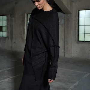 Könnte beinhalten: Ein schwarzer Langarmpullover mit sichtbaren Nahtdetails. Der Pullover wird mit einer schwarzen Hose kombiniert. Das Model steht in einem Betonraum mit Fenstern.