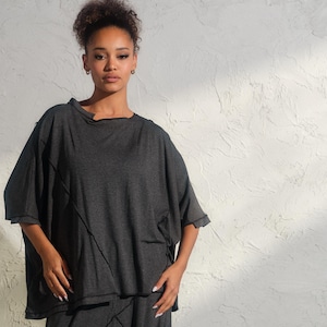 Può includere: Un top grigio antracite largo e morbido con un design leggermente asimmetrico, abbinato a pantaloni grigi antracite con un design geometrico unico. Il look è completato da stivaletti neri alla caviglia.