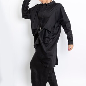 May include: A black long-sleeved shirt with a draped design and a black fedora hat. The shirt has a collar and a long pendant necklace. The person is wearing black pants and white shoes.