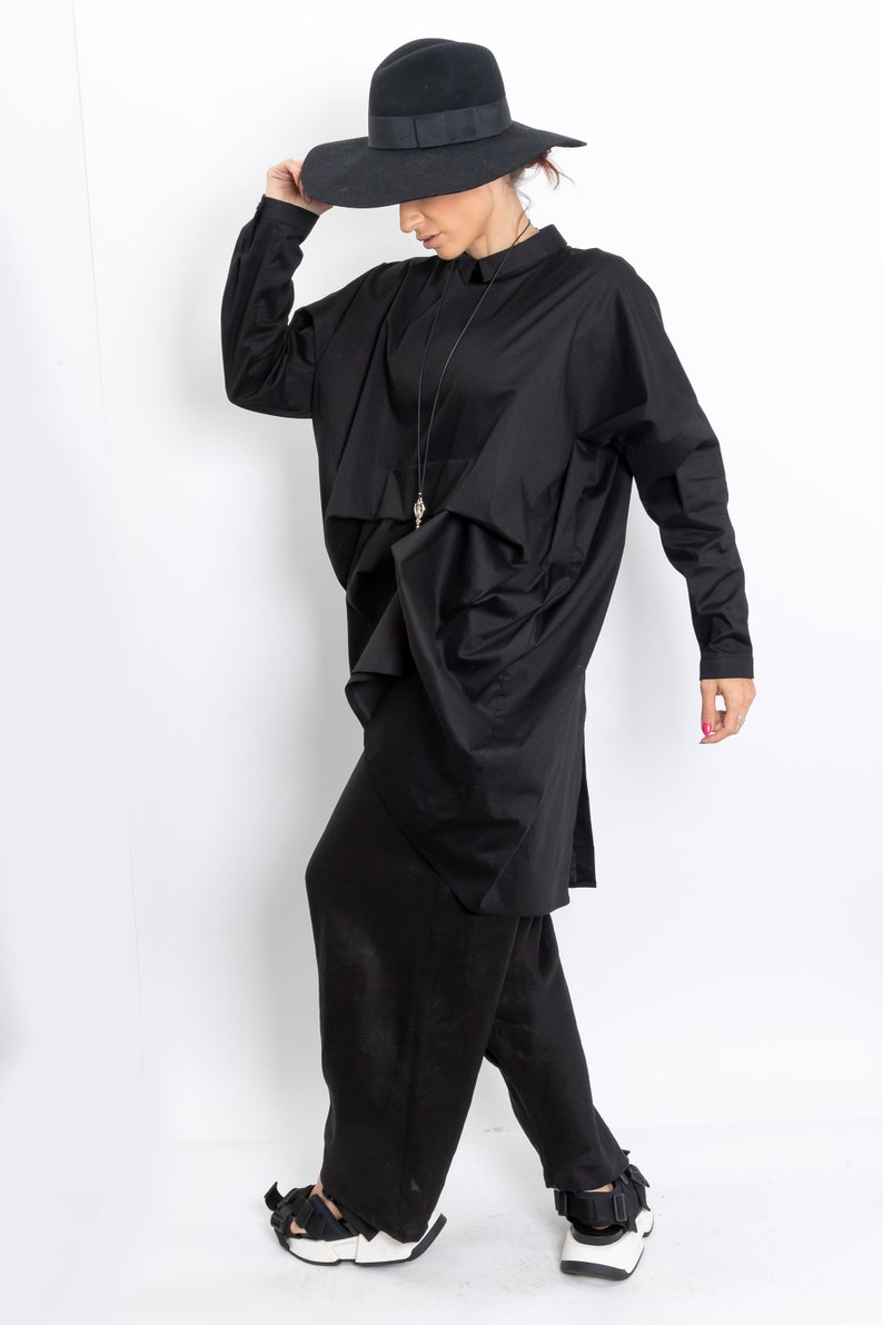 May include: A black long-sleeved shirt with a draped design and a black fedora hat. The shirt has a collar and a long pendant necklace. The person is wearing black pants and white shoes.
