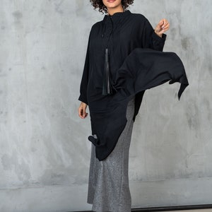 May include: A woman wearing a black long-sleeved shirt with a tassel necklace and a gray skirt. She is standing in front of a gray wall and is looking at the camera.