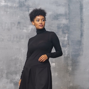 May include: A woman wearing a black turtleneck sweater and a black skirt. She is standing in front of a gray textured wall.