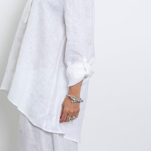 May include: A white linen shirt with long sleeves and a buttoned cuff. The shirt has a relaxed fit and a small collar. A silver bracelet with charms and rings are visible on the wrist.