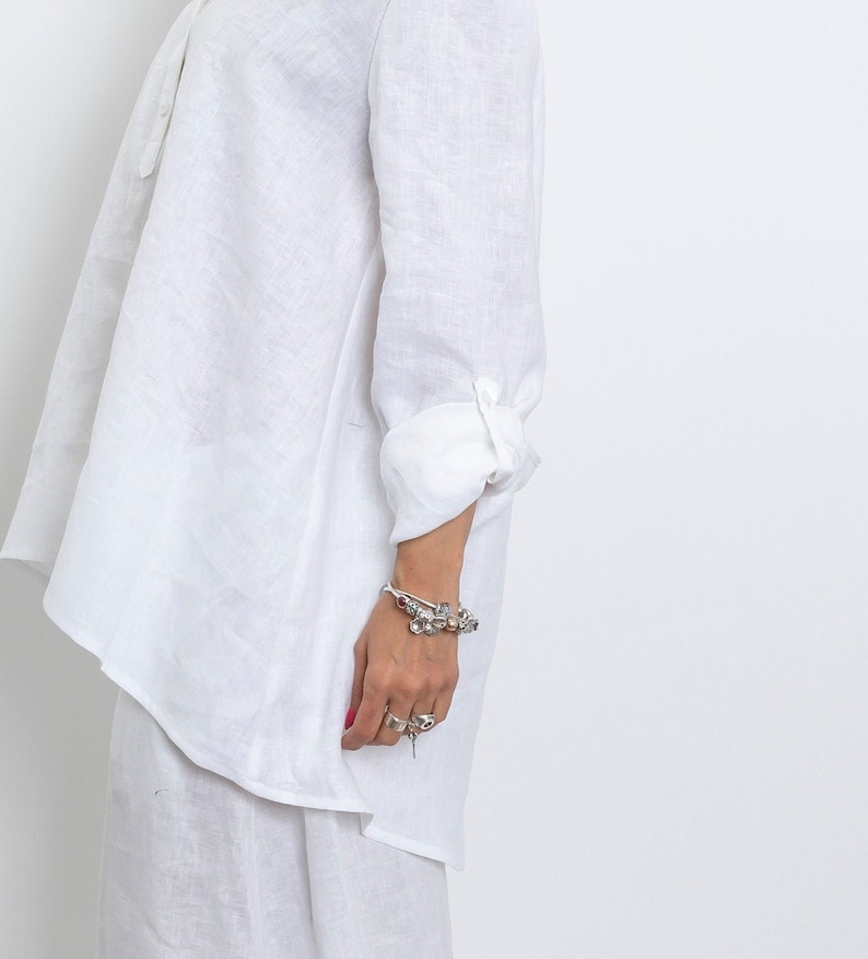 May include: A white linen shirt with long sleeves and a buttoned cuff. The shirt has a relaxed fit and a small collar. A silver bracelet with charms and rings are visible on the wrist.