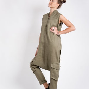 May include: A woman wearing a long, olive green linen jumpsuit with a sleeveless top and cargo pockets. She is standing with her right hand on her hip and looking up. She is wearing brown leather shoes.