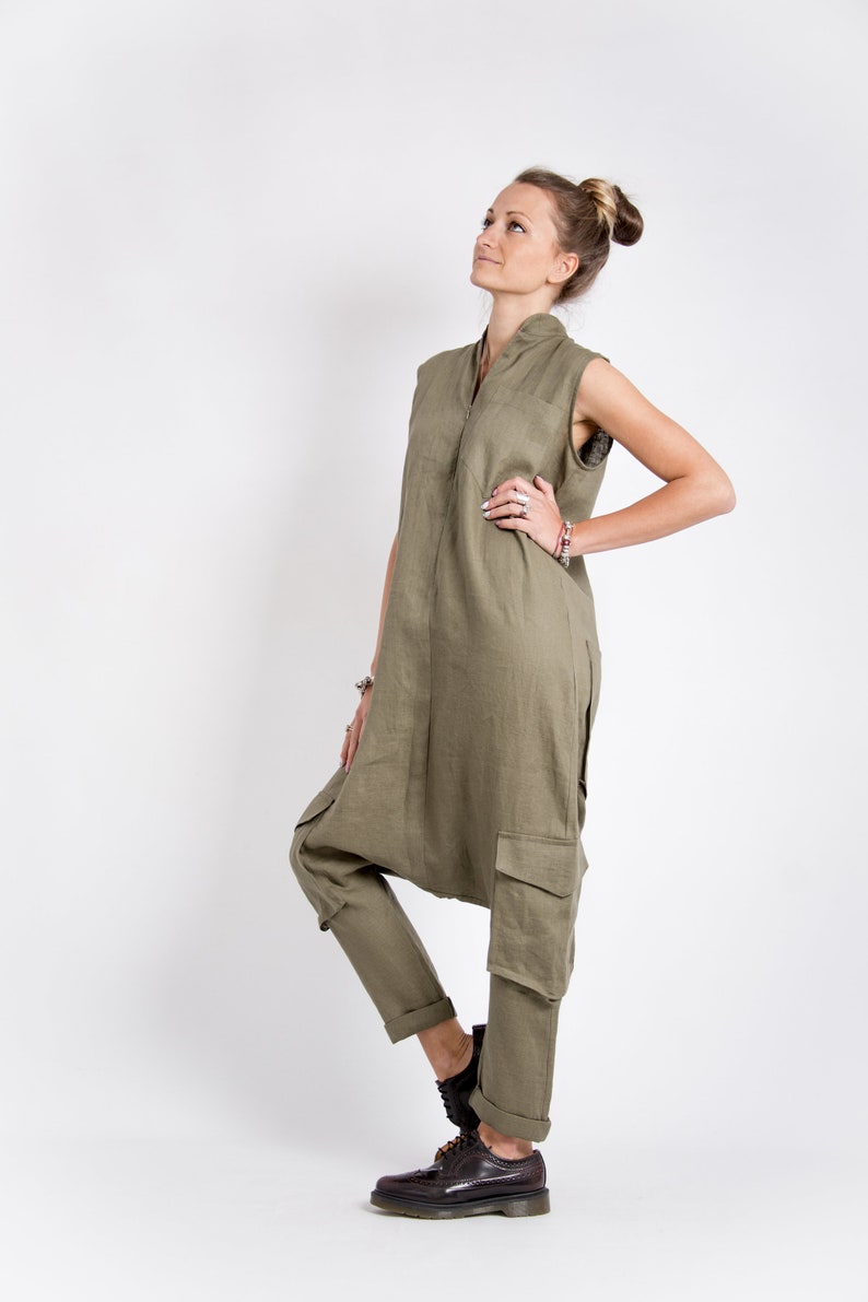 May include: A woman wearing a long, olive green linen jumpsuit with a sleeveless top and cargo pockets. She is standing with her right hand on her hip and looking up. She is wearing brown leather shoes.