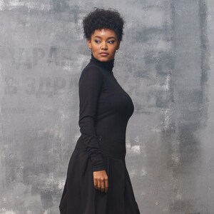 May include: A woman wearing a black turtleneck top and a black skirt. She is standing in front of a gray textured wall.