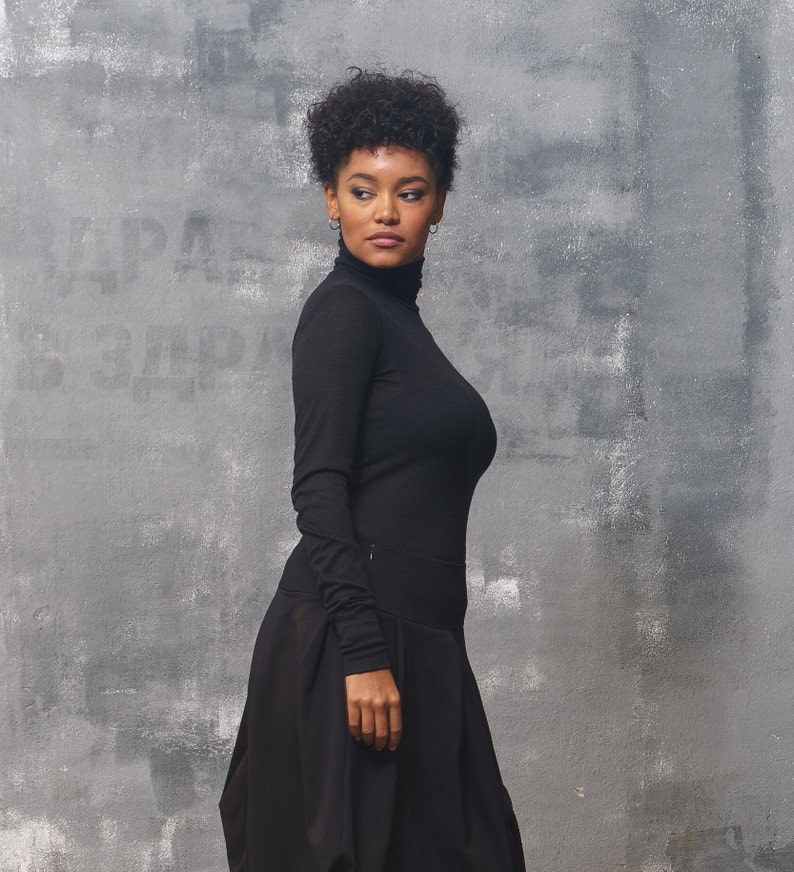 May include: A woman wearing a black turtleneck top and a black skirt. She is standing in front of a gray textured wall.