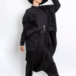 May include: A woman wearing a black, oversized shirt with a key pendant necklace and black pants. She is wearing a black hat and black sandals.