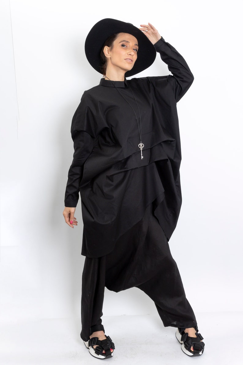 May include: A woman wearing a black, oversized shirt with a key pendant necklace and black pants. She is wearing a black hat and black sandals.