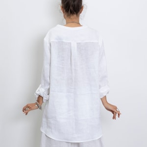 May include: White linen tunic shirt with long sleeves, rolled up at the elbows. The shirt has a simple collar and a slightly curved hem. The model is wearing a matching white skirt. The outfit is made of a lightweight, breathable fabric.