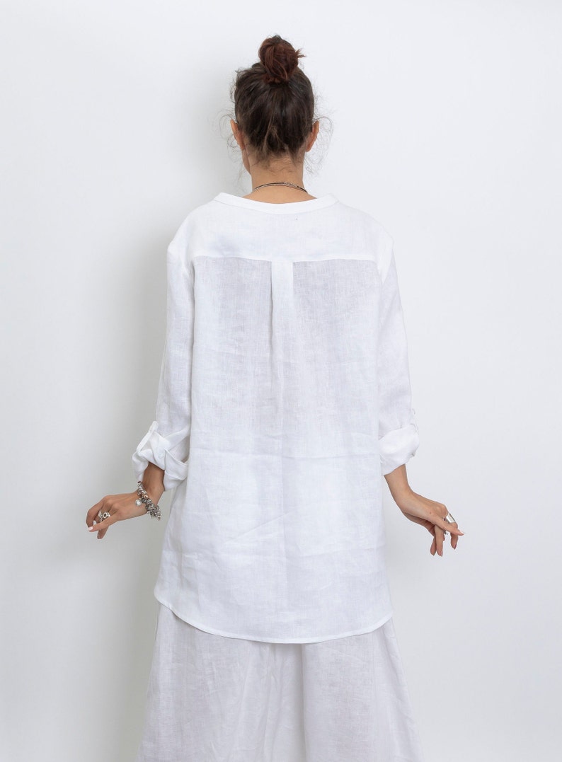 May include: White linen tunic shirt with long sleeves, rolled up at the elbows. The shirt has a simple collar and a slightly curved hem. The model is wearing a matching white skirt. The outfit is made of a lightweight, breathable fabric.