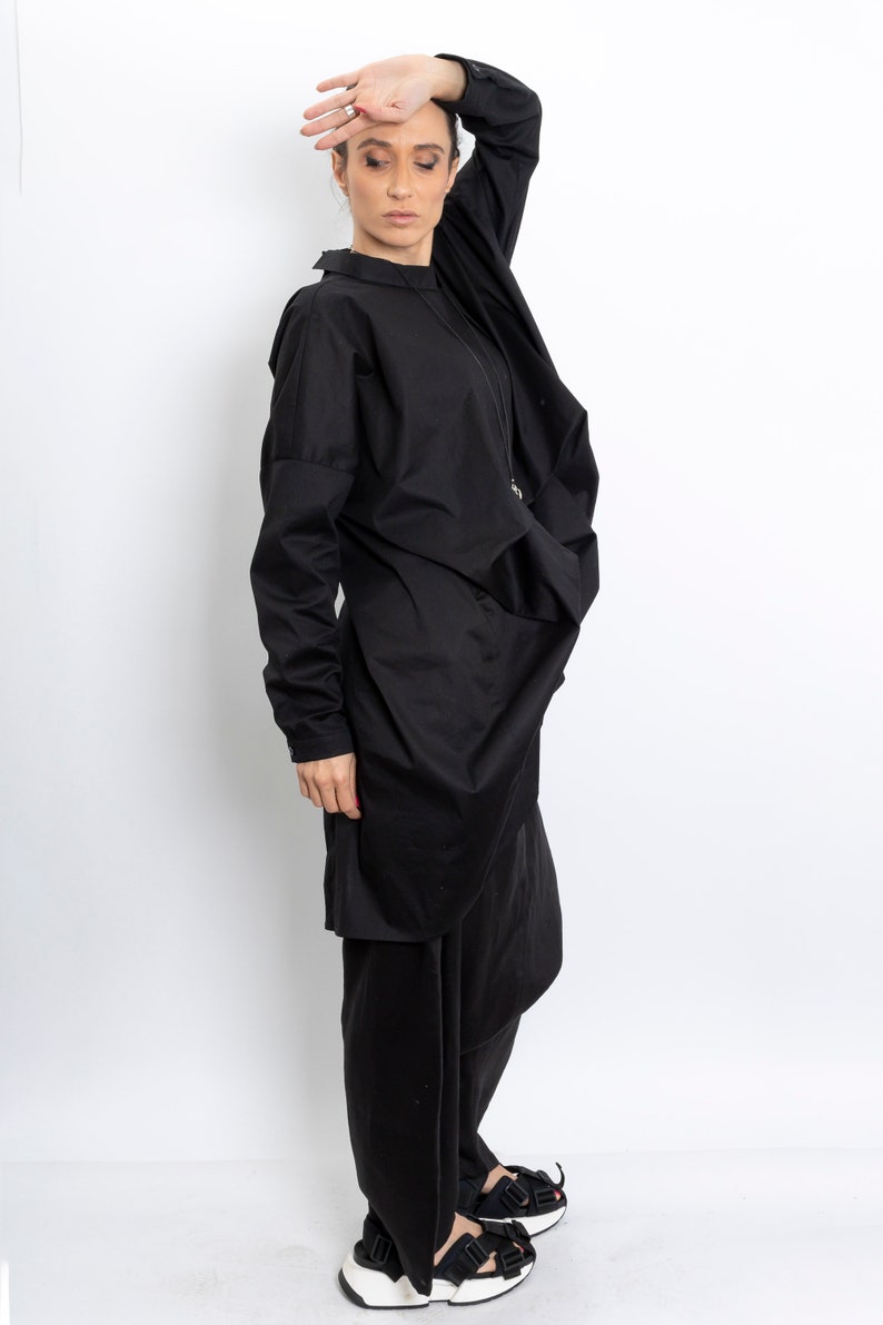 May include: A black long-sleeved shirt with a draped design and a black skirt. The person is wearing black sandals with white soles.