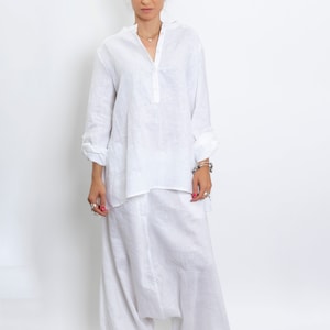 May include: A white linen outfit consisting of a long-sleeved tunic shirt with a mandarin collar and loose-fitting harem pants. The outfit is made of a lightweight, breathable fabric, perfect for warm weather. The model is wearing silver rings and bracelets.